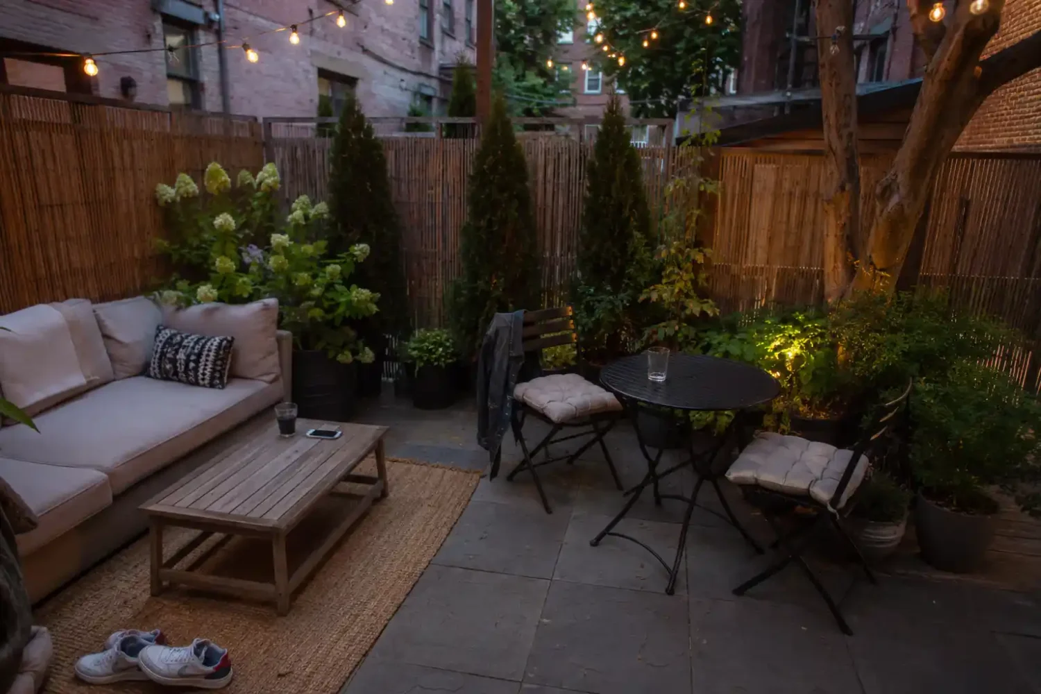 Small townhouse patio divided into a seating zone and dining zone with string lights, tall planters, and a bistro table at dusk.