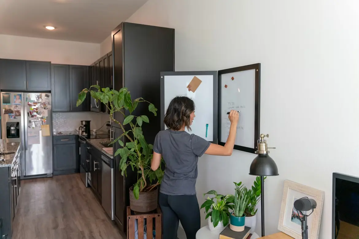 Woman writing on a wall-mounted whiteboard organizer in a modern kitchen with dark cabinets and plants.