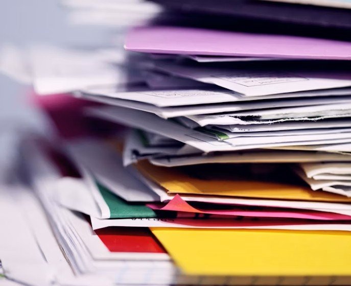 Messy stack of colorful paper, envelopes, and folders piled up on a desk.