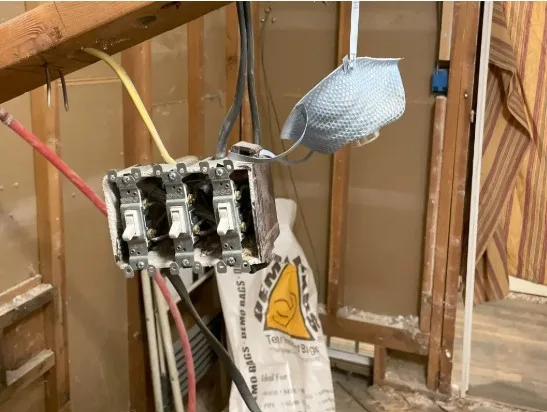 Close-up of exposed electrical wiring and a three-switch box hanging loose during a bathroom remodel, with wires visible and a dust mask hanging nearby. Construction debris and framing are seen in the background.