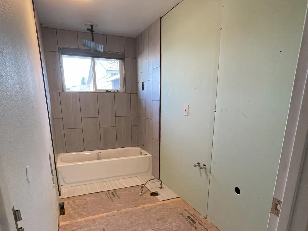 Bathroom remodel with a new bathtub installed under a window, gray tile partially finished on the shower wall, and green board drywall on the side wall ready for finishing. Subfloor is exposed with plumbing fixtures visible.