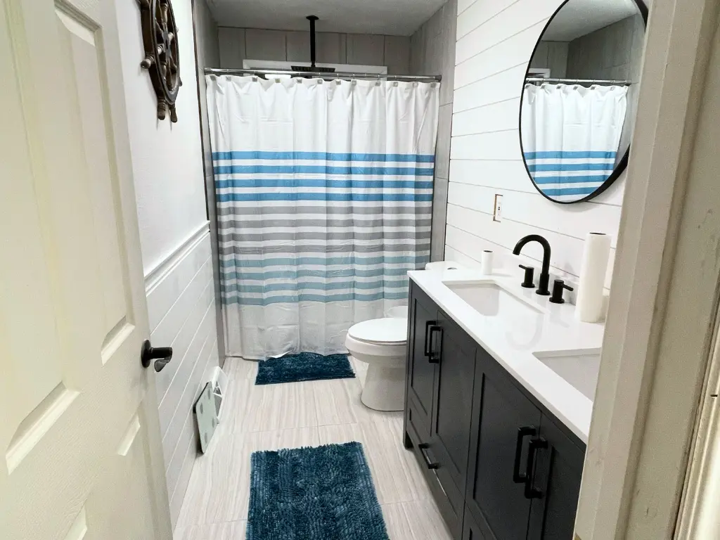 Finished bathroom with white shiplap walls, a black vanity with double sinks, round mirror, and matte black fixtures. A striped blue and gray shower curtain and teal bath rugs add a coastal pop of color.