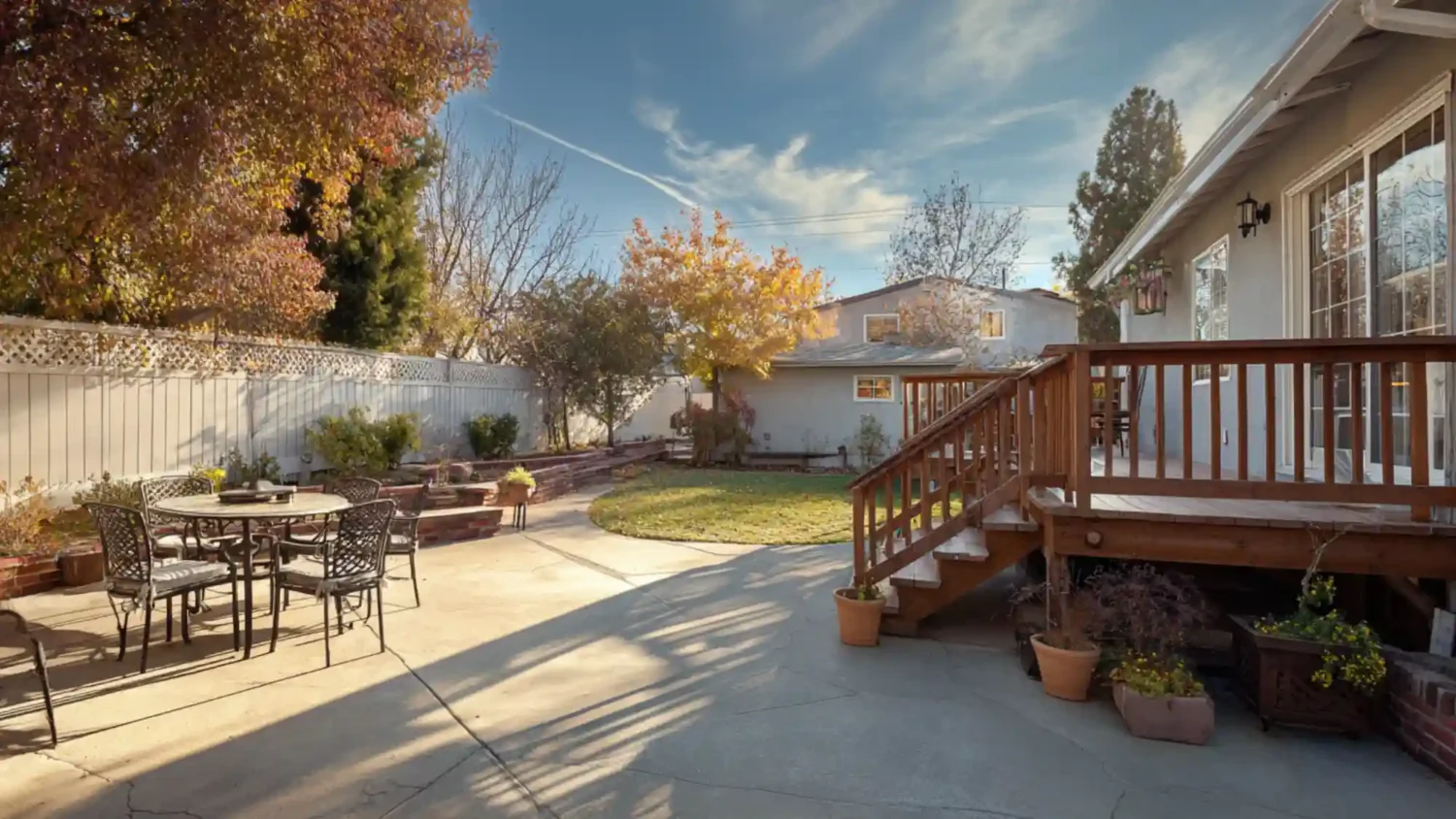Patio vs deck shown in a real backyard, concrete patio with dining set on the left and an elevated wood deck with stairs on the right, fall landscaping.