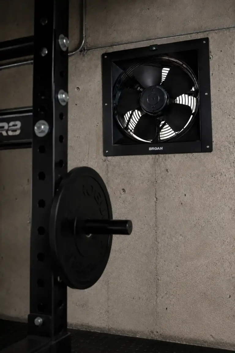 Garage gym ventilation Broan wall mounted exhaust fan on concrete wall with power rack and water bottle.