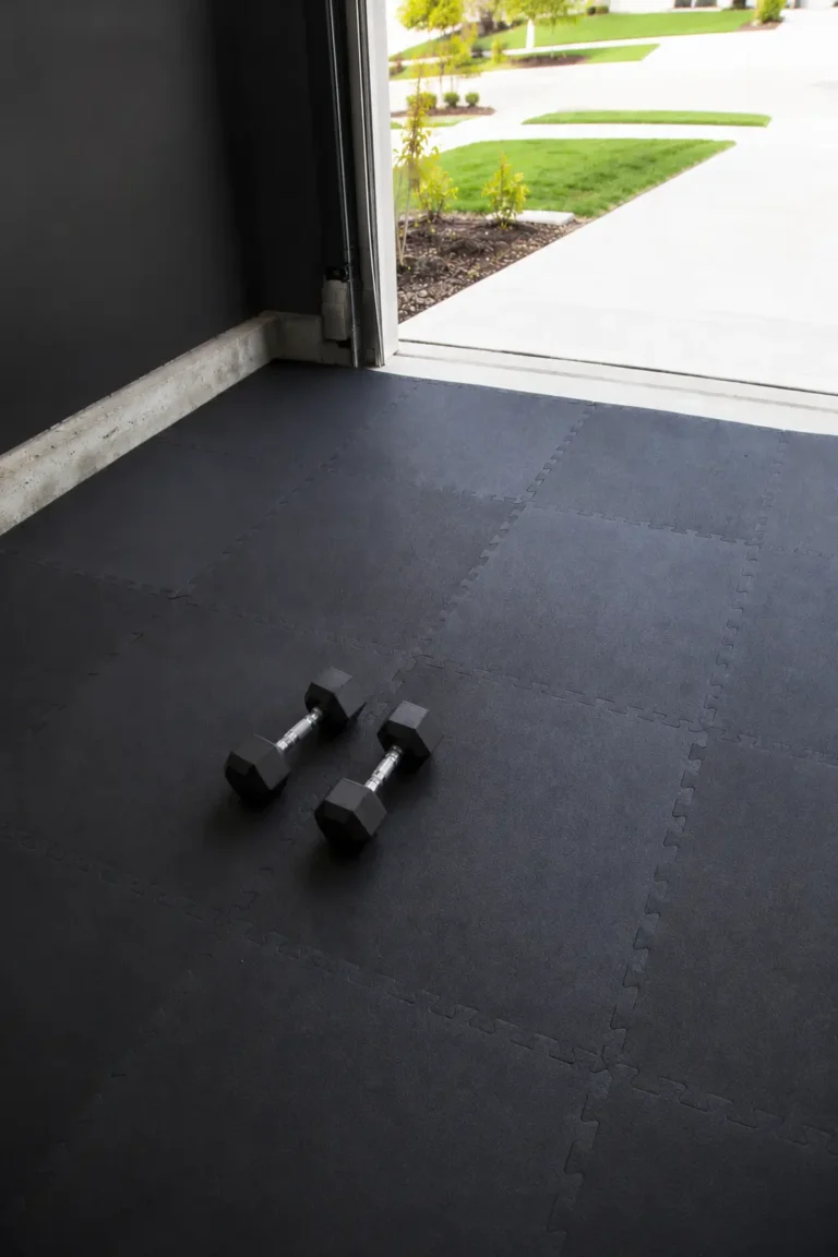 Garage gym flooring with black interlocking rubber mats and dumbbells in open garage.
