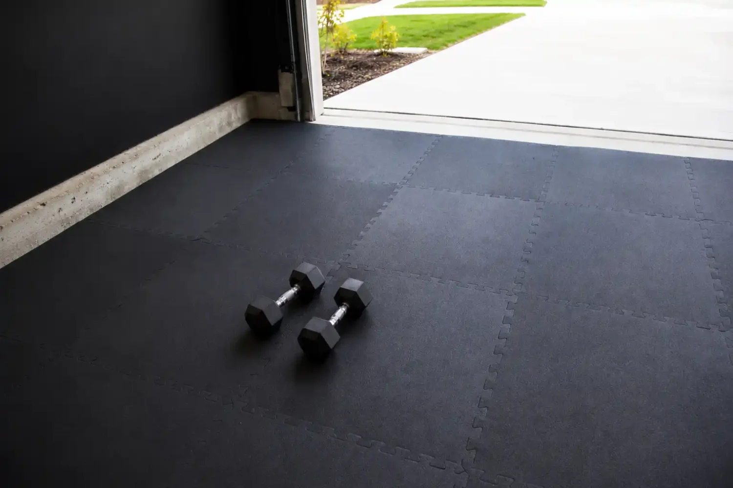 Black interlocking rubber gym mats laid on garage gym flooring with natural light and dumbbells.