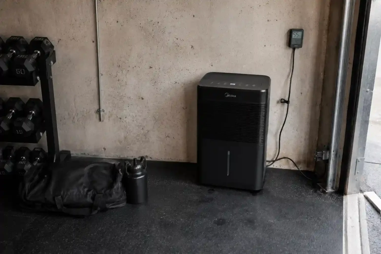 Dehumidifier and hygrometer mounted on garage gym wall next to a dumbbell rack to control humidity.