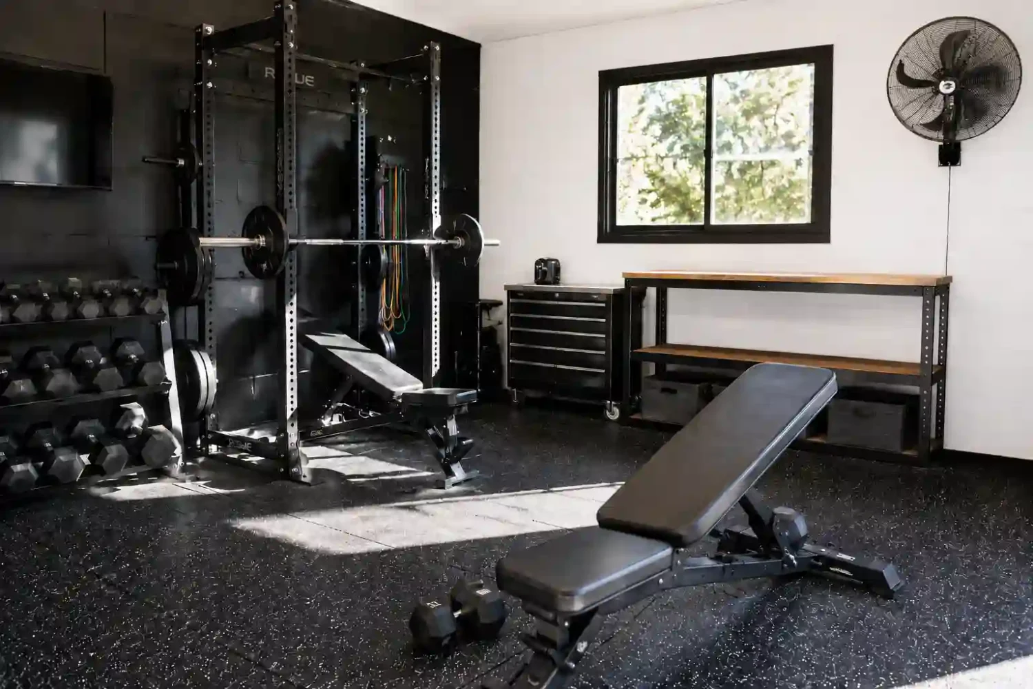 Garage gym ventilation setup with wall mounted fan, power rack, dumbbells, and rubber flooring in a home gym.