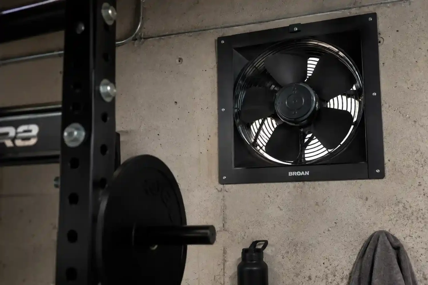Garage vent fan mounted high on a concrete wall next to a power rack in a black garage gym.