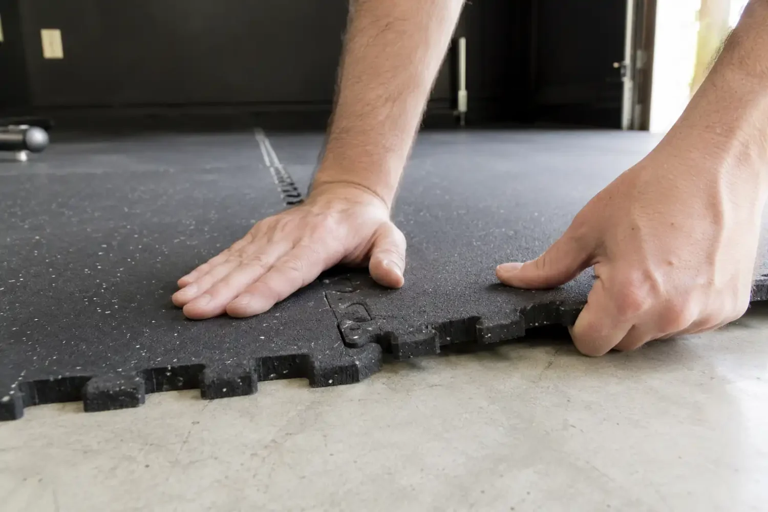 Hands installing interlocking rubber gym mat tiles on concrete garage floor.