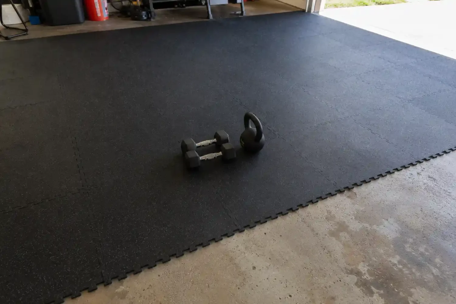 Dark rubber interlocking gym mats installed over a stained concrete garage floor with dumbbells and a kettlebell on top.