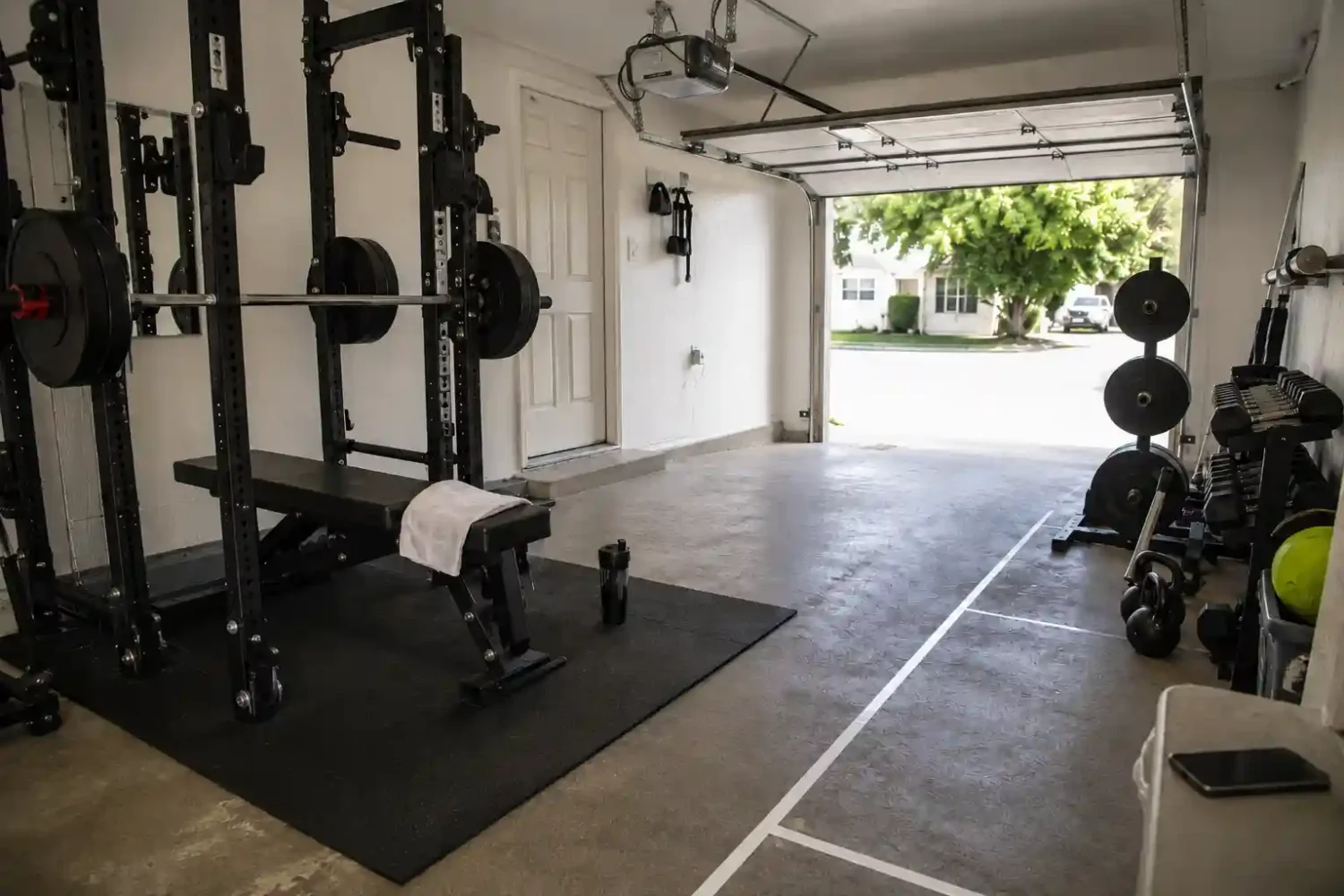 Garage gym storage ideas with weight plates on a rack-mounted post, a lifting belt and wrist wraps hanging on wall hooks, and a power rack against a white garage wall.