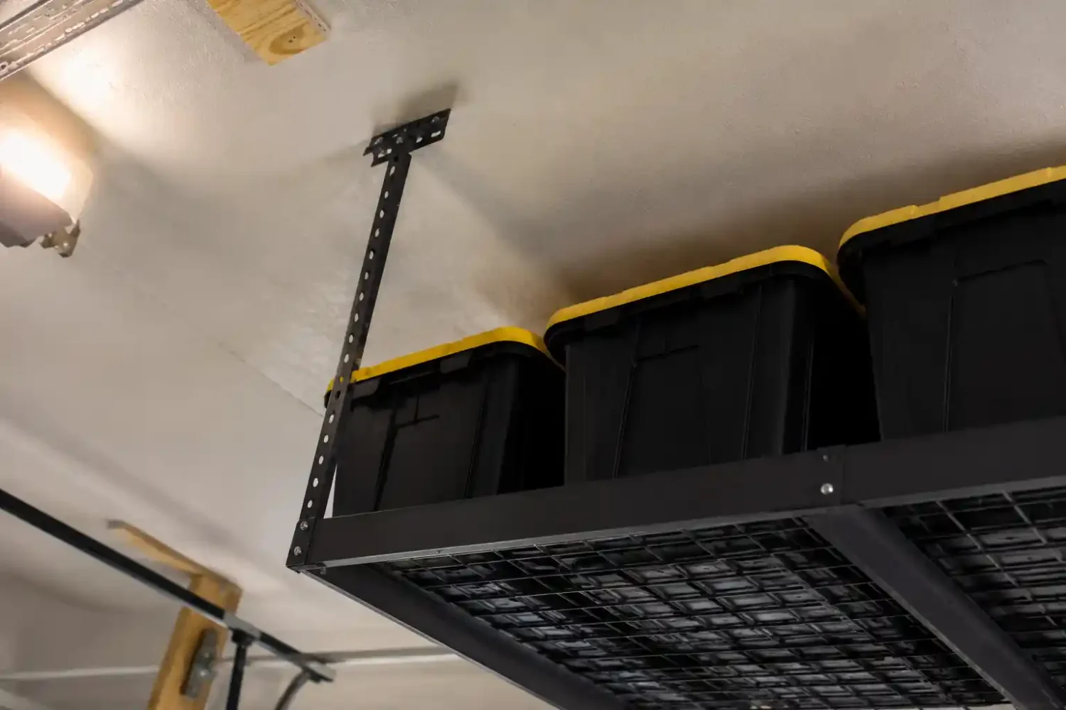Overhead garage storage rack viewed from below at an angle, holding black storage totes with yellow lids, mounted near a garage light fixture and ceiling framing.