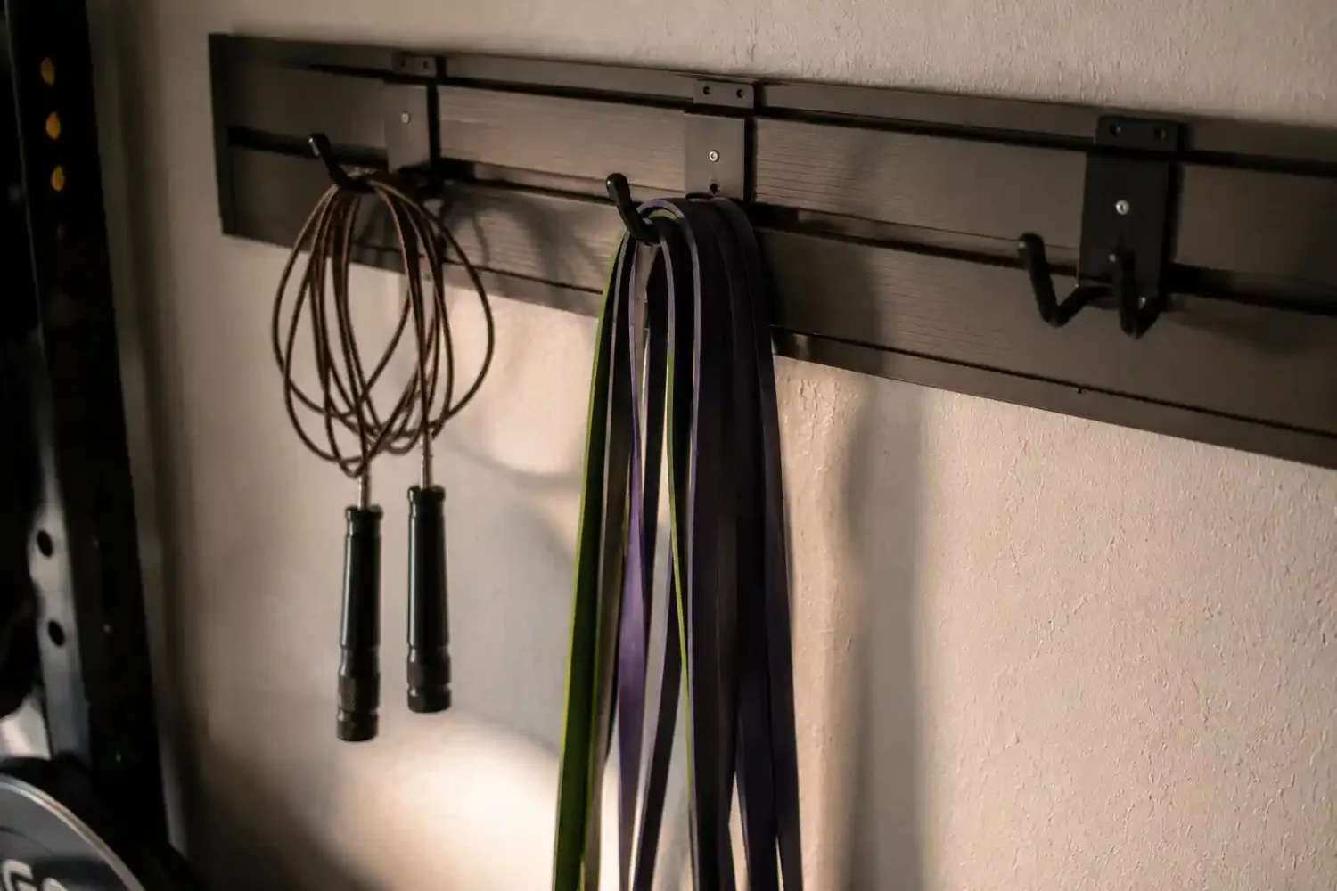 Speed rope and resistance bands hanging on slatwall hooks in a garage gym, with empty hooks visible on a light textured wall.