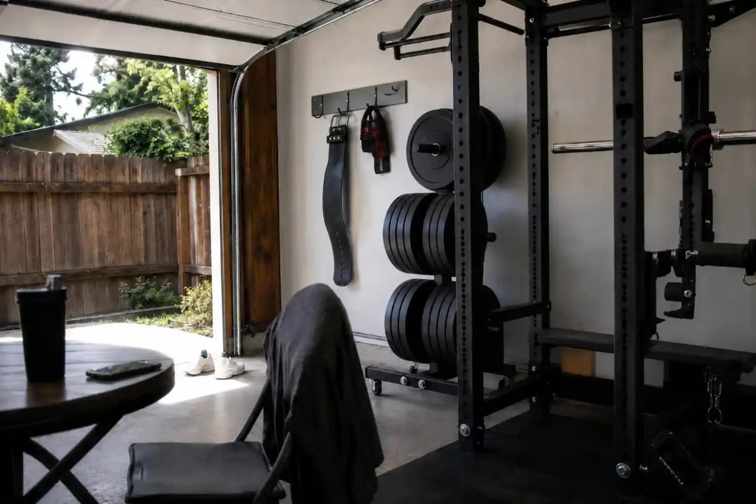 Garage gym storage ideas with weight plates on a rack-mounted post, a lifting belt and wrist wraps hanging on wall hooks, and a power rack against a white garage wall.