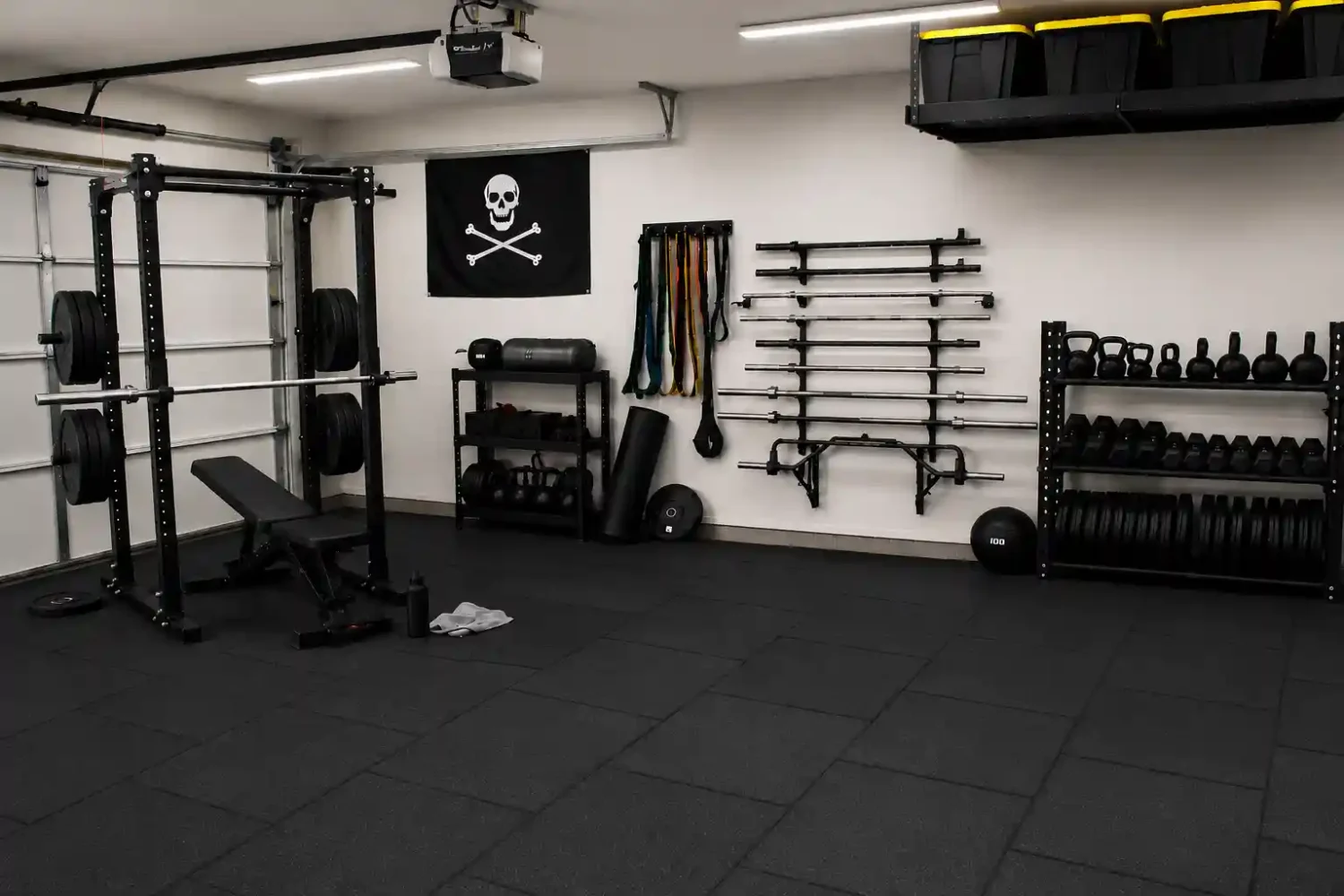 Garage gym with distinct zones, a power rack and bench in the training area, and kettlebell and dumbbell racks along the back wall.